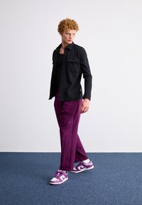 Svart långärmad skjorta med knappar och två bröstfickor, i kombination med avslappnade lila byxor och lila- och vitfärgade sneakers.