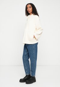 Witte fleece jas met een ritssluiting bij de kraag en twee voorzakken, gecombineerd met blauwe spijkerbroek en zwarte platformlaarzen.