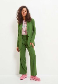 Groene op maat gemaakte pak met brede broekspijpen en een blazer. Daaronder een lichtroze blouse met bloemenpatronen. Chunky roze platform schoenen.