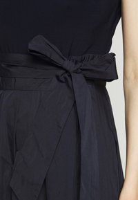 En svart tygbälte knutet i en rosett runt midjan på ett mörkt plagg, som visar en del av en arm på höger sida.