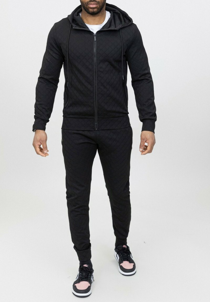 Survêtement matelassé noir comprenant un hoodie zippé, un pantalon slim et des cordons de serrage. Assorti à des baskets noires et roses.