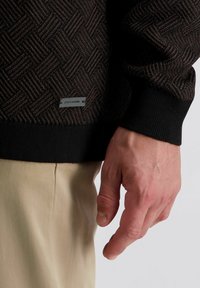 Schwarzer Pullover mit Fischgrätenmuster und gerippten Bündchen. Verfügt über ein elegantes graues Etikett an der unteren Vorderseite.