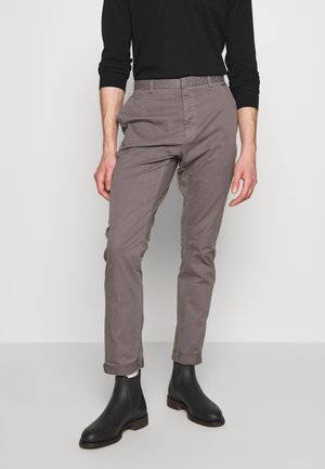 Mężczyzna ubrany w dopasowane szare spodnie podwinięte przy nogawce, czarną bluzkę z długim rękawem, białe skarpetki i czarne buty Chelsea, stojący na jednolitym tle.