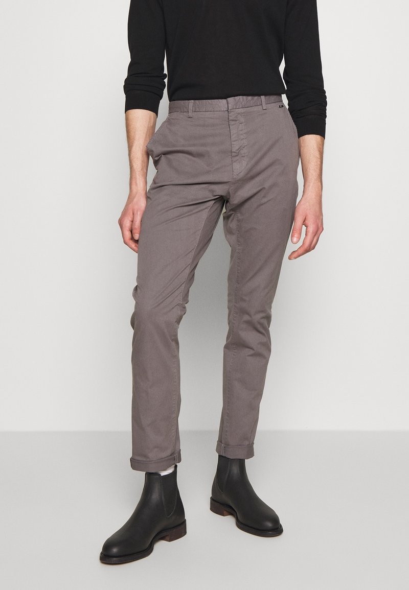 Homme portant un pantalon gris ajusté retroussé à l'ourlet, un haut noir à manches longues, des chaussettes blanches et des bottines Chelsea noires, debout devant un fond uni.