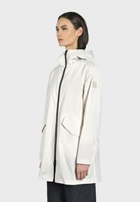 Giacca impermeabile bianca con cappuccio, zip frontale, tasche laterali e polsini elastici. Presenta un tessuto texturizzato e un logo discreto sulla manica.