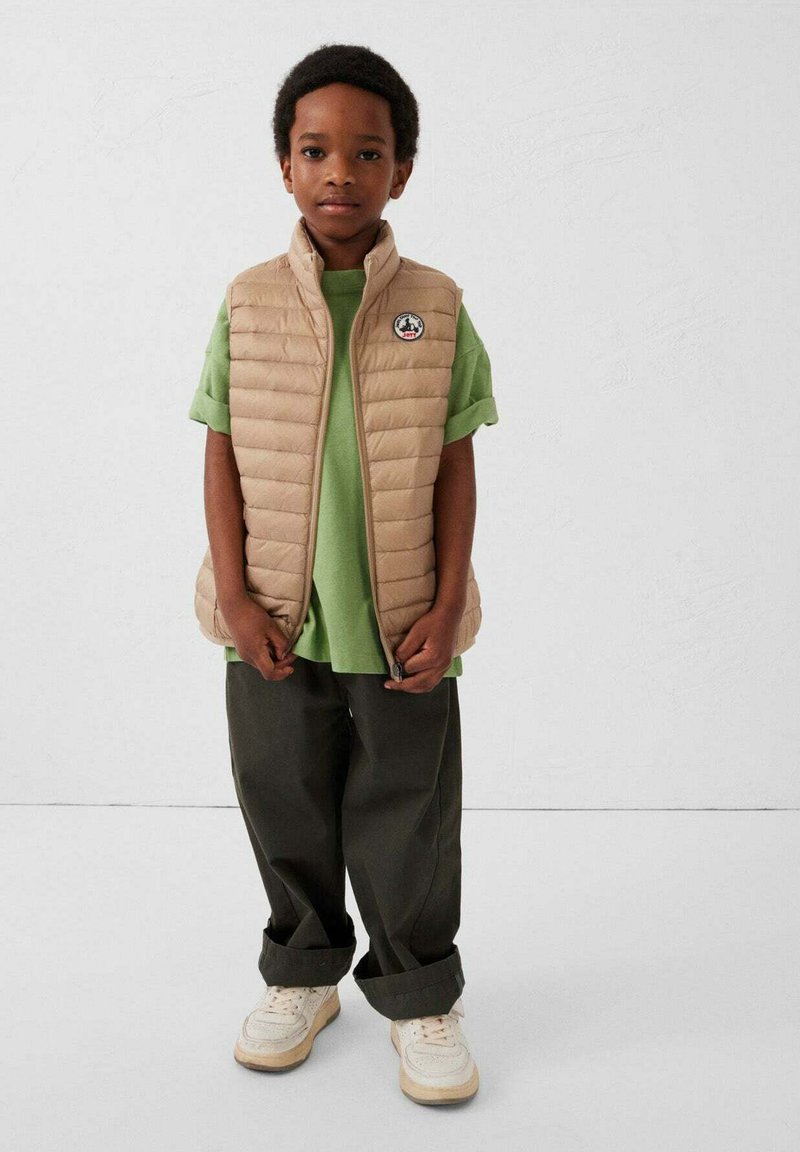 Beige gewatteerd vest over een groen T-shirt, gecombineerd met donkere losse broek en crèmekleurige sneakers. Heeft een logo op het vest. Effen achtergrond.