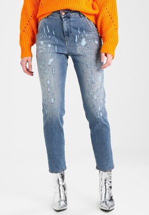 Osoba ubrana w jasnoniebieskie podniszczone dżinsy, jaskrawo pomarańczowy sweter z dzianiny oraz błyszczące srebrne botki, stojąca na białym tle.