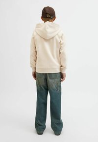Jasno beżowy hoodie z kapturem na sznurku, w połączeniu z wyblakłymi niebieskimi spodniami o szerokich nogawkach z detalem kieszeni. Widok z tyłu.