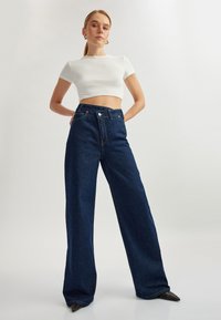 Jeans de mezclilla oscura de pierna ancha y cintura alta, con una textura suave, cierre de botón metálico y bolsillos frontales, combinados con un top corto blanco ajustado.
