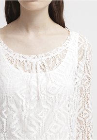 Bílé krajkové blůzky s jemnými geometrickými vzory, kulatým výstřihem a dlouhými, průhlednými rukávy. Měkká textura a lehký design.