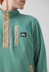 Pullover in pile di colore verde menta con colletto alto, pattina con bottoni beige e patch del logo nero. Texture morbida con un design casual.