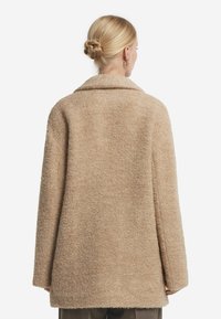 Beige, oversize rock med ett strukturerat, fluffigt material. Har en stor krage och vida ärmar, vilket ger en avslappnad passform.