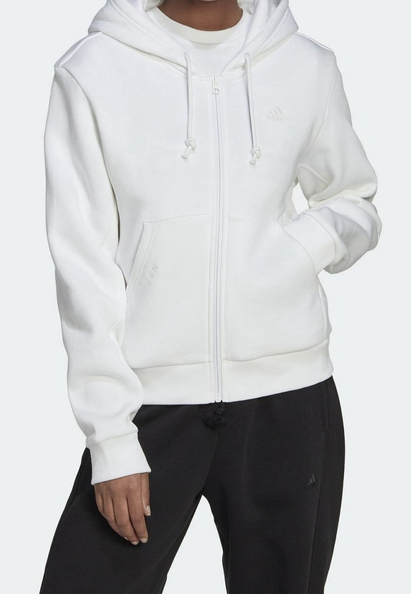 Haut à zip blanc en tissu doux, avec une poche avant, des cordons de serrage et des poignets côtelés. Porté par-dessus une chemise blanche et un pantalon noir.