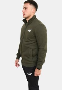 Giacca a zip verde oliva con colletto alto, polsini a coste e tasche laterali. Presenta il logo Puma bianco sul petto. Texture liscia.