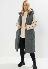 Long gilet matelassé gris avec capuche, présentant un motif en matelassage et une fermeture éclair. Associé à un pull crème et un pantalon noir, agrémenté de bottes noires.