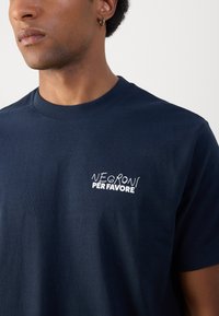 Marineblå bomull T-skjorte med hvit brodert tekst "NEGRONI PER FAVORE" på venstre bryst. Korte ermer og rund hals.