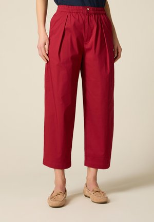 Kvinde iført højtaljede, løstsiddende røde bukser med plisseringer og beige mokkasiner, stående foran en ensartet lys baggrund.