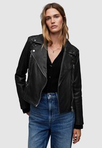 Giubbotto in pelle nera da motociclista con dettagli trapuntati sulle spalle, cerniere argentate e silhouette aderente, abbinato a un maglione nero e jeans a vita alta.