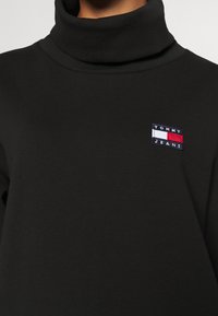 Pull à col roulé noir en coton doux, avec un petit logo rectangulaire Tommy Jeans en rouge, blanc et bleu sur le côté gauche supérieur.