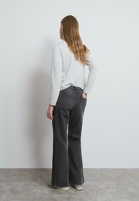 Mujer con cabello largo de espaldas a la pared, vistiendo una camiseta de manga larga gris claro, jeans negros de pierna ancha con lados estampados y zapatillas deportivas.