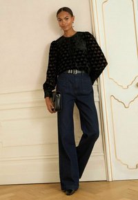 Melna zīda blūze ar sirds rakstiem, pūkainām piedurknēm. Augstās jostasvietas tumšas džinsu bikses. Modelis tur mazu melnu somiņu. Vienkāršs fons.