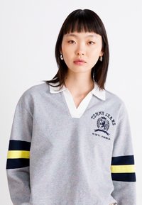 Maglione grigio con colletto bianco, caratterizzato da un logo blu e la scritta "TOMMY JEANS EST. 1985," con strisce navy e gialle sulle maniche.