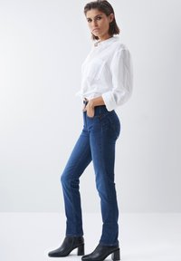 Jeans de tiro alto azules de denim, combinados con una camisa blanca de botones y botines negros de tacón, de pie sobre un fondo sencillo.