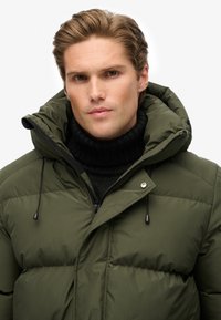 Olivenfarvet pufferjakke med høj krave, lynlåslukning foran og snoretræk. Båret over en sort ribbet turtleneck sweater. Glat tekstur.