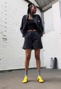 Zwarte textuur crop jack en bijpassende shorts, gecombineerd met een zwart tanktop. Felgele sportschoenen en grijze sokken. Minimalistische achtergrond.