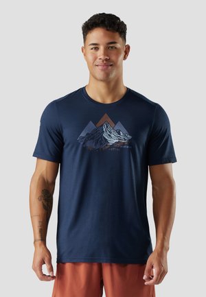 Ung mand iført en mørkeblå t-shirt med et bjerghajlk og rustfarvede shorts, stående mod en ensfarvet lys baggrund.