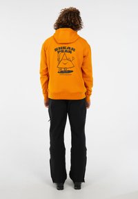 Person mit lockigem Haar, der einen orangefarbenen Hoodie mit Berggrafik und schwarze Hosen trägt, steht mit dem Rücken zu einem weißen Hintergrund.