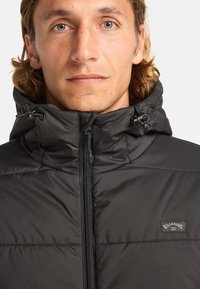 Giacca a vento nera con colletto alto e coulisse regolabile. Presenta una chiusura con zip frontale e un piccolo patch logo Billabong.