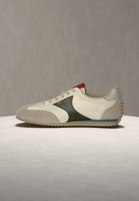 Baskets basses avec des panneaux en cuir beige, blanc et noir, détail rouge sur la languette et semelle en caoutchouc rainurée sur une surface neutre.
