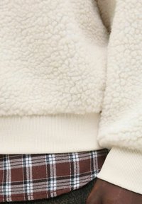 Crèmekleurige fleece pullover met een gestructureerd oppervlak, voorzien van een ribbelige crèmekleurige zoom. Daaronder een bruin geruit shirt met zwart-witte accenten.