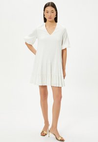 Robe blanche courte avec un décolleté en V, des manches ballon et un ourlet à volants en tissu texturé. Le mannequin porte des sandales à talons dorées et des boucles d'oreilles.