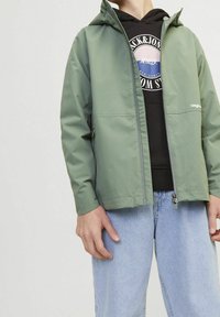 Osoba w zielonej kurtce z zamkiem błyskawicznym założonej na czarną bluzę z grafiką, zestawiona z jasnoniebieskimi dżinsami na jednolitym tle.