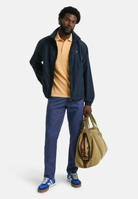 Marineblå vindtät jacka, ljust orange polo, blå byxor, blå sneakers med vita detaljer. Bär en beige duffelväska med bruna detaljer.