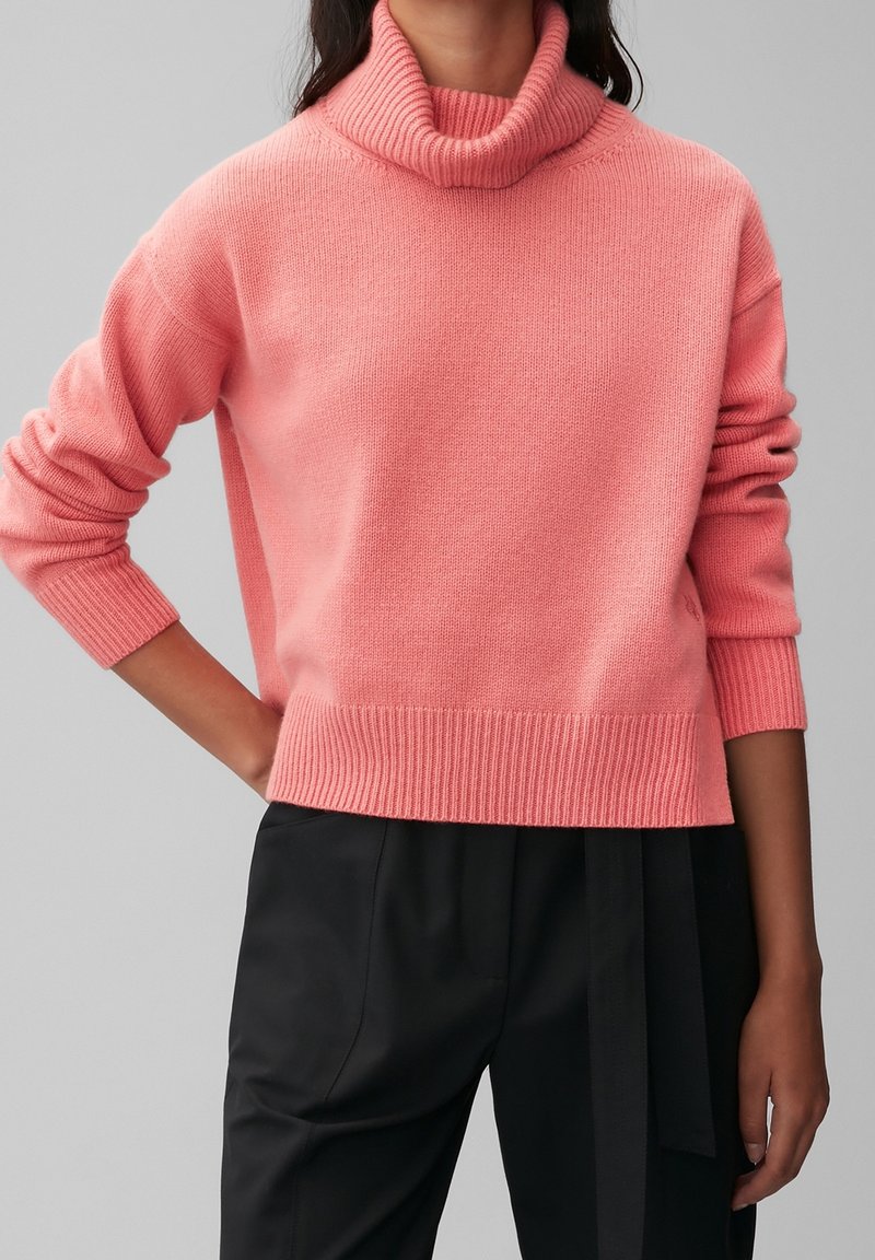 Woman wearing a pink turtleneck sweater with ribbed cuffs and hem, paired with black high-waisted trousers with a fabric belt.