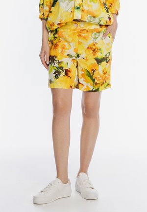 Osoba ubrana w żółte szorty w kwiaty i pasującą koszulę, w białych trampkach, stojąca na tle jednolitego białego tła.