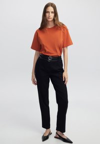 T-shirt arancione a maniche corte abbinato a pantaloni neri a vita alta con una cintura e una fibbia lucida discreta, completato da scarpe nere slip-on.