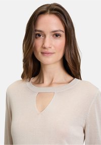 Tops en maille beige à manches longues avec un col rond et une découpe en forme de clé. Texture douce et design ajusté. Coutures décoratives visibles.