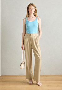 Light blue tank top with decorative shoulder straps, paired with high-waisted beige trousers. Model holding a small beige bag, standing on wooden floor.