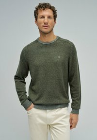 Zielony sweter z dzianiny, z okrągłym dekoltem, teksturowanym materiałem oraz ściągaczami przy mankietach i u dołu. Dyskretne logo na lewej piersi. W zestawieniu z jasnymi spodniami.