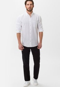 White button-up shirt with a chest pocket, rolled-up sleeves, paired with black pants and white sneakers, on a neutral background.