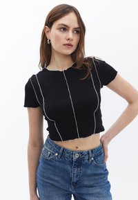 Schwarzes, kurzes T-Shirt mit kurzen Ärmeln, ausgestattet mit kontrastierenden weißen Nähten und einem gewellten Ausschnitt. Kombiniert mit blauen Jeans.