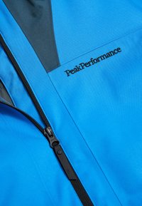 Blaue Outdoor-Jacke mit schwarzen Akzenten, ausgestattet mit einem vorderen Reißverschluss, glatter Textur und PeakPerformance-Logo auf der Brust.