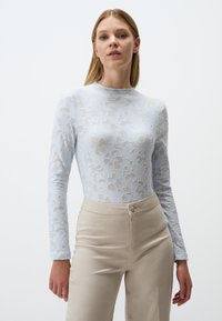 Top a maniche lunghe in pizzo blu chiaro con motivo floreale, colletto alto e silhouette aderente, abbinato a pantaloni beige a vita alta.