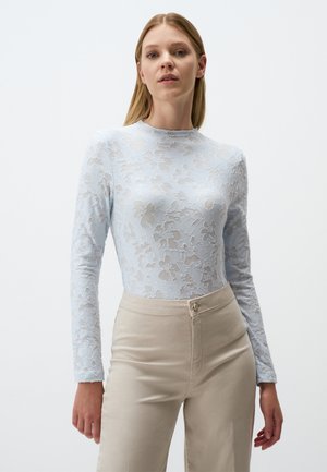 Top a maniche lunghe in pizzo blu chiaro con motivo floreale, colletto alto e silhouette aderente, abbinato a pantaloni beige a vita alta.