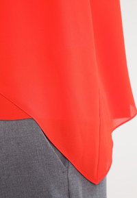 Top rosso brillante con orlo asimmetrico abbinato a pantaloni in tessuto grigi, che mostrano texture e sovrapposizione del capo in vita.