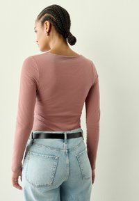 Langärmliges, enganliegendes Hemd in staubigem Rosé, kombiniert mit hochgeschnittenen hellblauen Jeans und einem schwarzen Gürtel, der einen lässigen Look präsentiert.